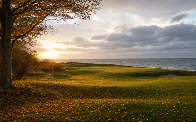 Herbstliches Fairway an der Küste mit Laub, tief stehender Sonne und wehender Fahne am Grün